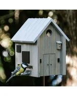 Kohlmeise an der Futteröffnung des Multiholk Modern Nordic von Wildlife Garden, einem grauen Vogelhaus mit hellem Satteldach auf einem Pfosten im Garten.