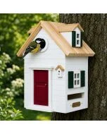 Kohlmeise am runden Einflugloch des Multiholk New England von Wildlife Garden, einem weißen Vogelhaus mit roter Tür, grünen Fensterläden und hellem Holzdach an einem Baumstamm.