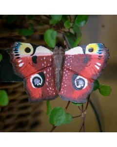 DecoButterfly Tagpfauenauge aus Holz von Wildlife Garden mit rotbraunen Flügeln, vier auffälligen Augenflecken und dunklen Flügelrändern zwischen grünen Blättern.