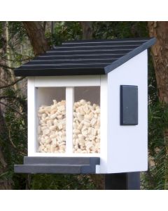 Eichhörnchen Futterhaus Weiß Ekorrmatare von Wildlife Garden auf einem Pfosten zwischen Bäumen, mit schwarzem Schrägdach und hellen Futterstückchen hinter den Sichtfenstern.