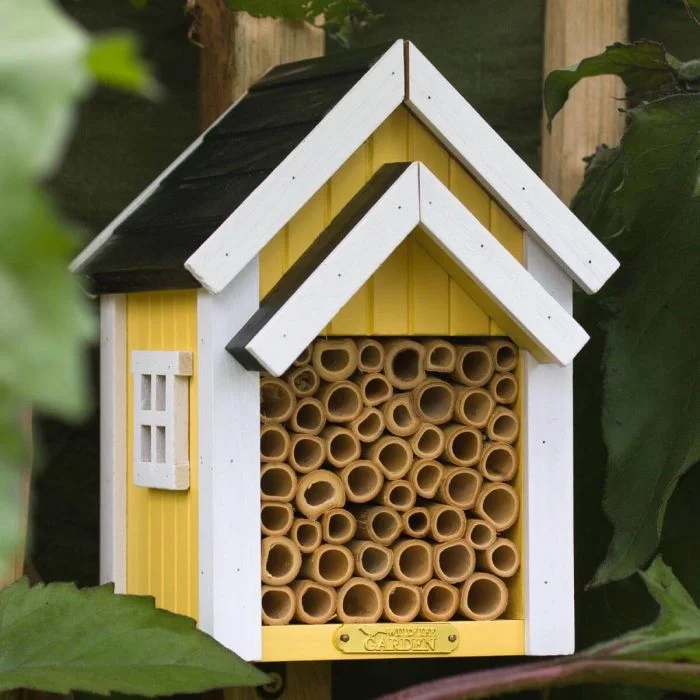 Bienenhotel Biholk Gelb von Wildlife Garden zwischen grünen Blättern im Garten, mit gelber Holzfassade, weißen Dachleisten und hohlen Bambusröhrchen.