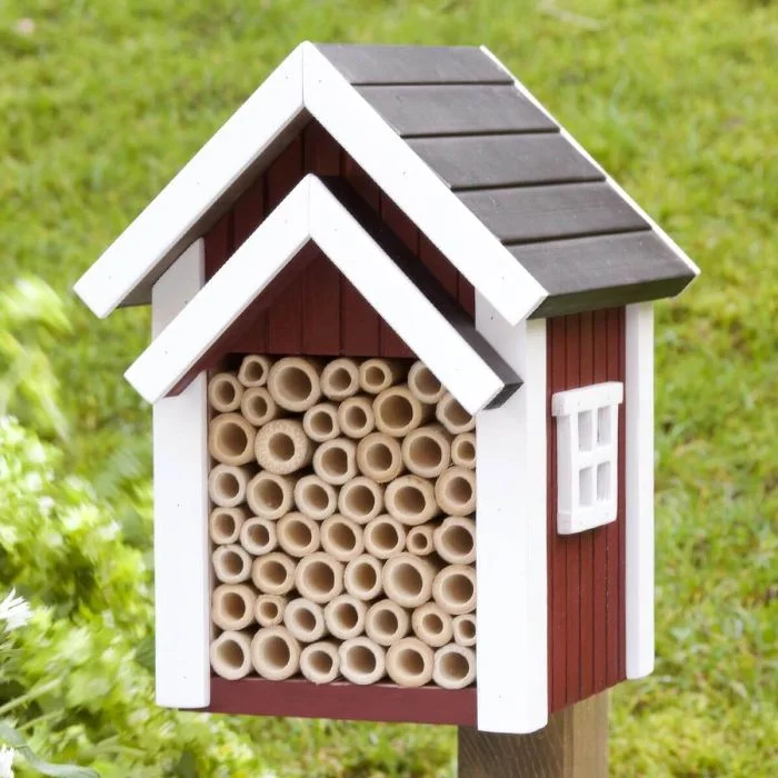 Bienenhotel Biholk Rot von Wildlife Garden auf einem Holzpfosten im grünen Garten, mit roter Holzfassade, weißen Dachleisten und hohlen Niströhren.