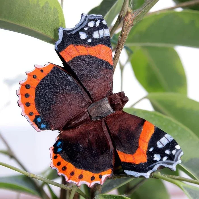 DecoButterfly Admiral von Wildlife Garden als Holzschmetterling zwischen grünen Blättern, mit dunkelbraunen Flügeln, orangefarbenen Binden und weißen Flecken.