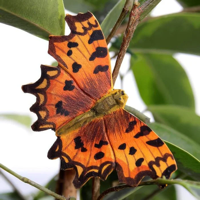 DecoButterfly C-Falter aus Holz von Wildlife Garden mit orangefarbenen Flügeln, schwarzen Flecken und gezackten Flügelrändern zwischen grünen Pflanzenblättern.