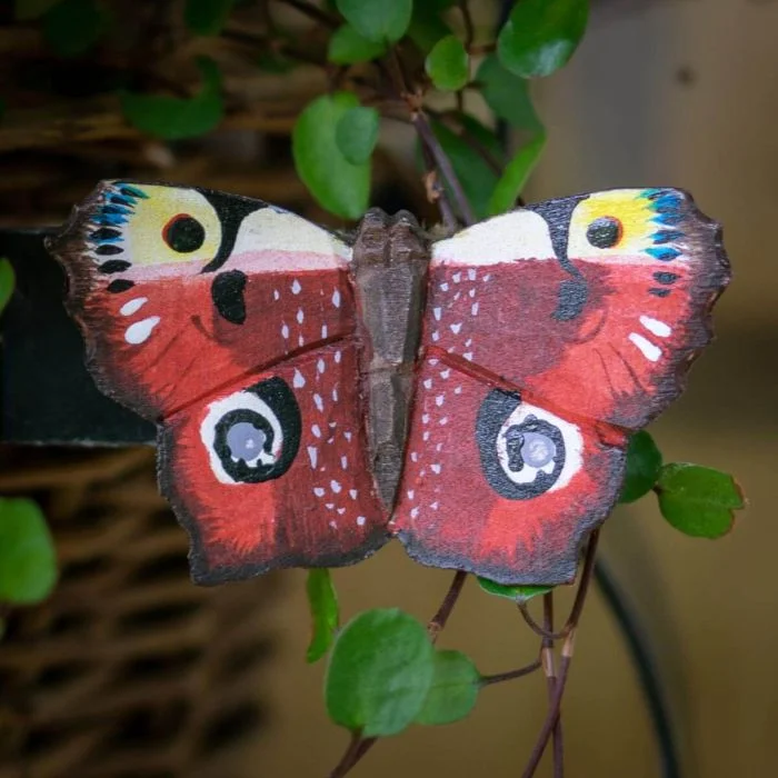 DecoButterfly Tagpfauenauge aus Holz von Wildlife Garden mit rotbraunen Flügeln, vier auffälligen Augenflecken und dunklen Flügelrändern zwischen grünen Blättern.