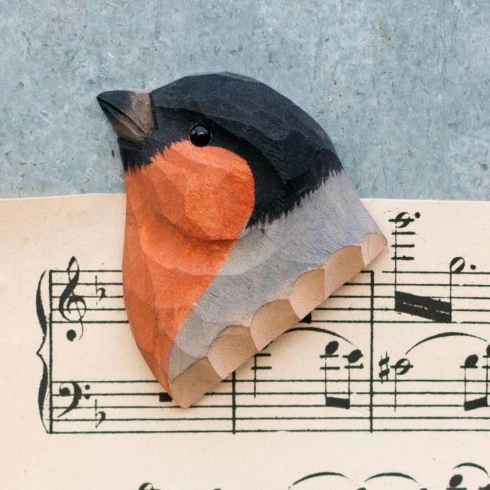 Kühlschrankmagnet Gimpel aus Holz von Wildlife Garden mit schwarzem Kopf, orange-roter Brust und grauem Flügel auf einem Notenblatt.