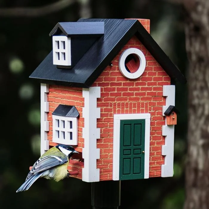 Kohlmeise an der Futteröffnung des Multiholk Brick Cottage von Wildlife Garden, einem Vogelhaus in roter Backsteinoptik mit grüner Tür und schwarzem Satteldach.